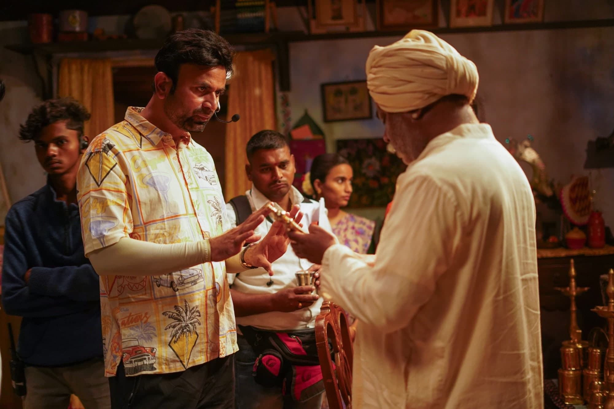 Santosh Davakhar, wearing a headset microphone, directs an elderly actor in a white turban on an interior set, with crew members gathered around them.