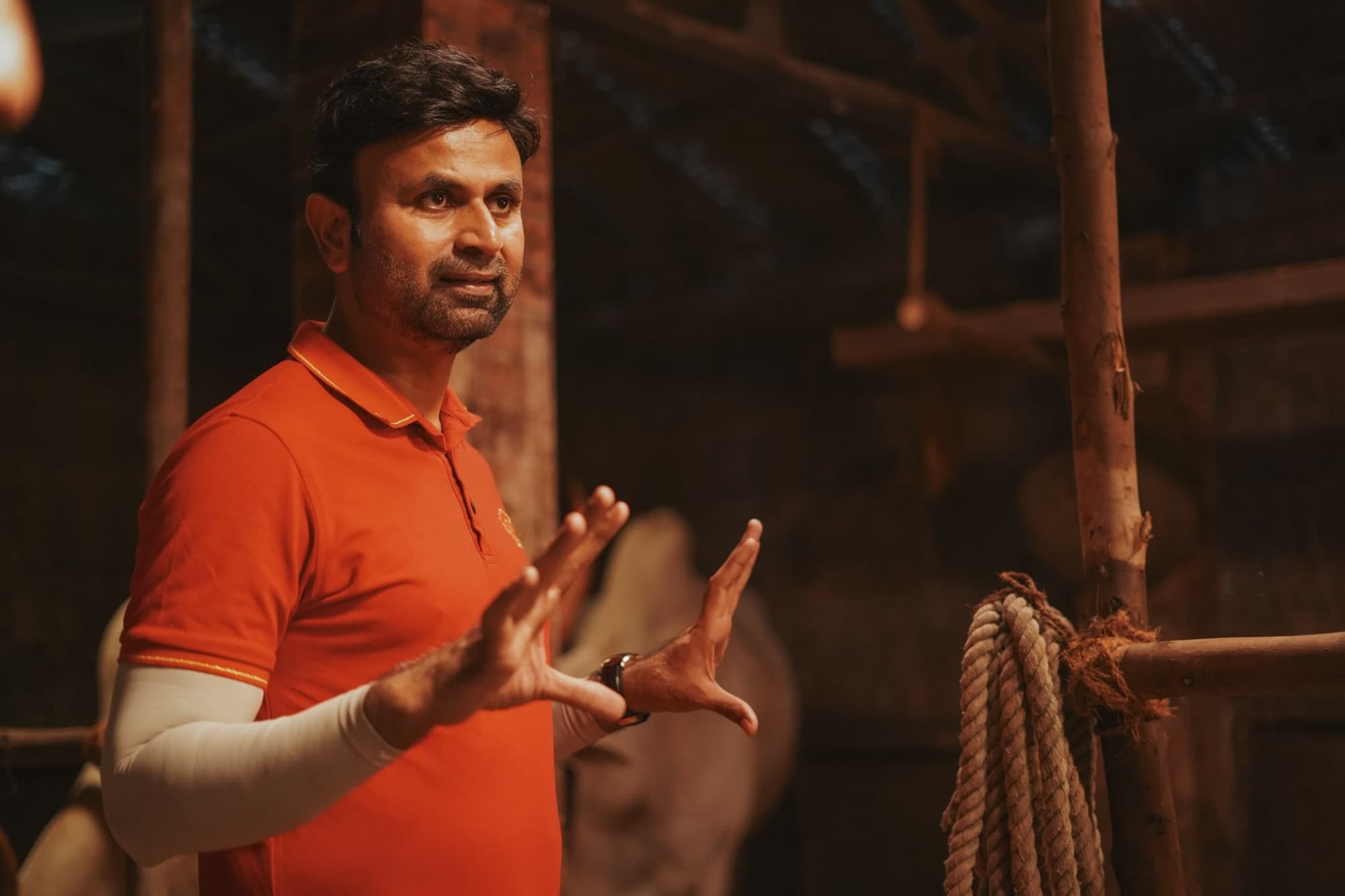 Santosh Davakhar, in an orange polo shirt, gestures with both hands as he directs on a dimly lit rustic barn set, a coil of rope hanging from a wooden post beside him.