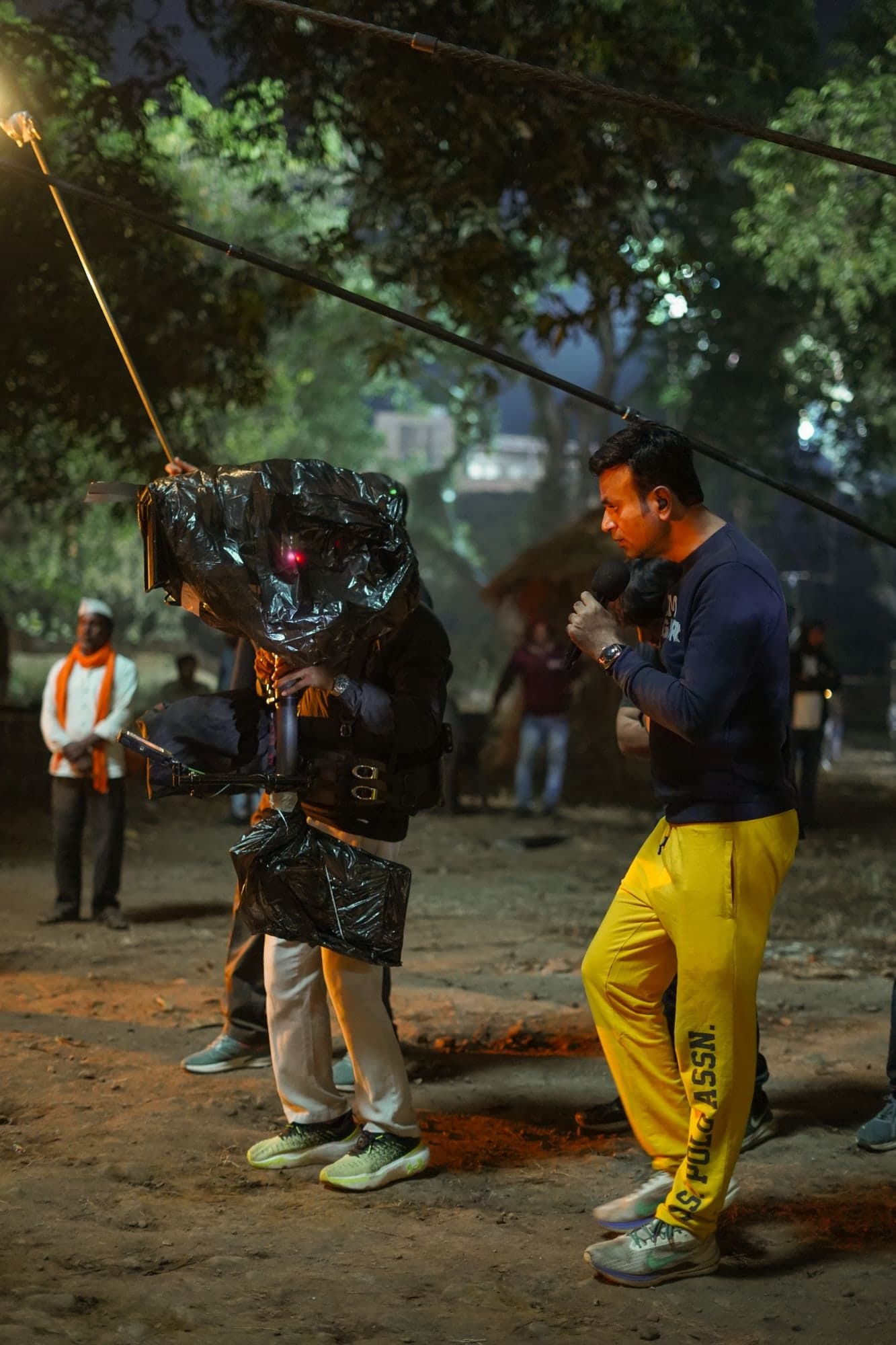 During a night shoot outdoors, Santosh Davakhar speaks into a microphone beside a camera rig wrapped in a plastic rain cover.