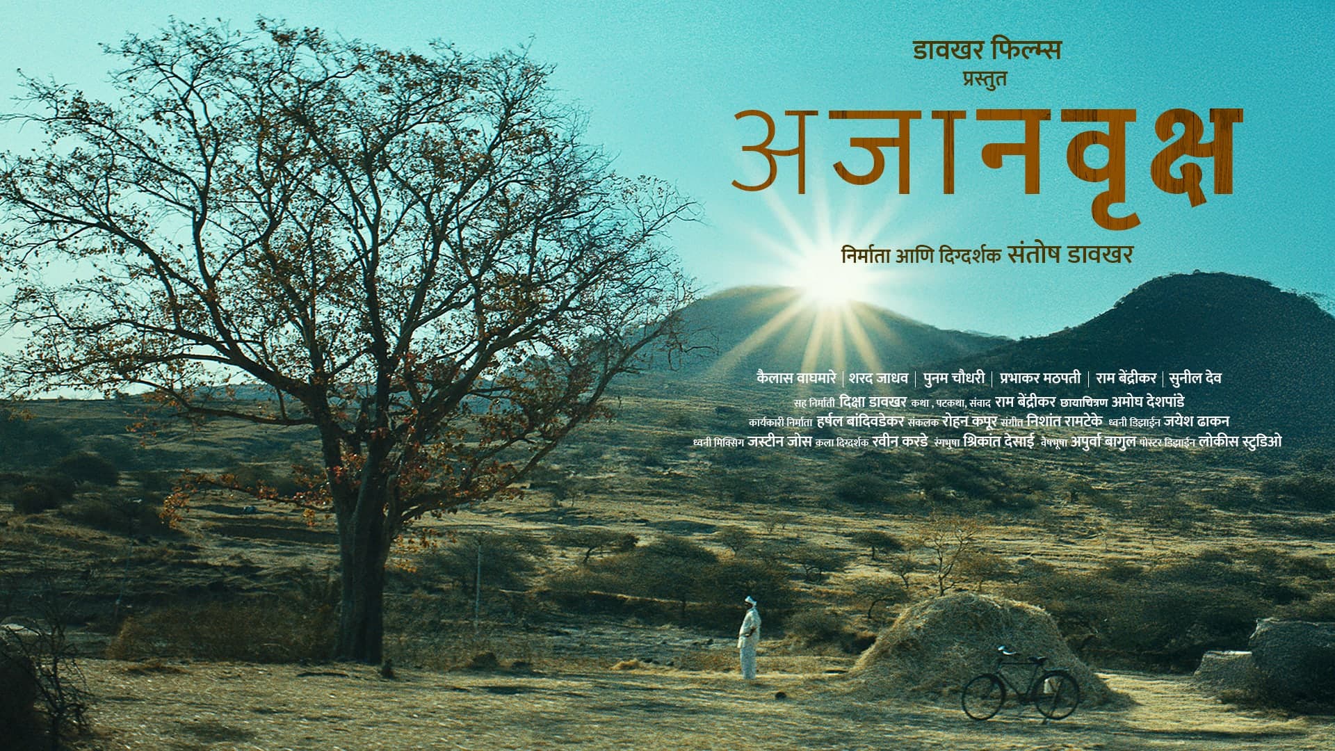 Ajanvruksh key art: a bare tree in a dry, hilly landscape as the sun bursts over a hill; a figure in white stands near a haystack and a bicycle. Text in Marathi: Davakhar Films presents; the title Ajanvruksh (अजानवृक्ष); producer and director Santosh Davakhar; billed names and the film’s credits.