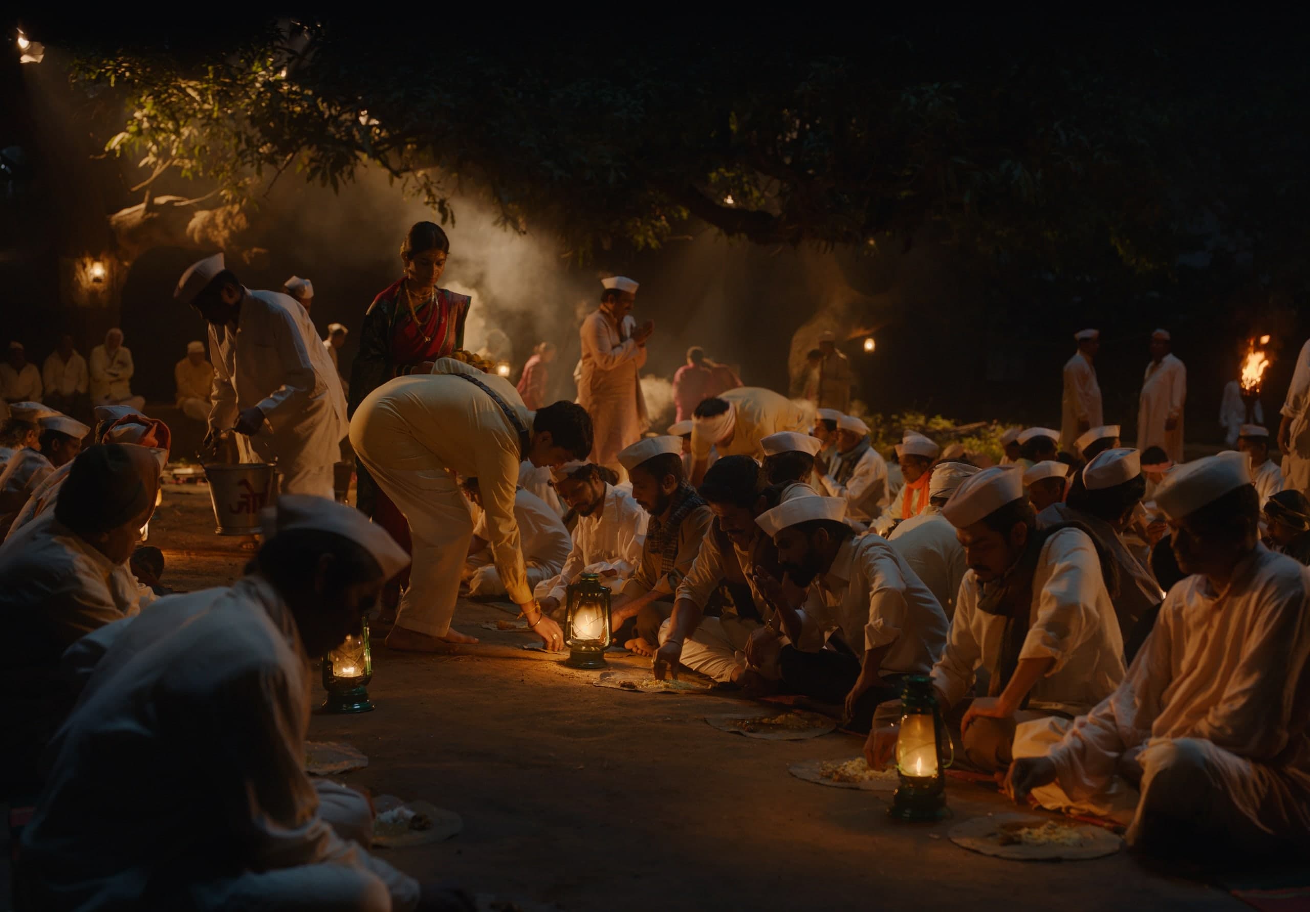 Men in white kurtas and Gandhi caps sit in rows on the ground, eating from leaf plates by the light of green lanterns, while food is served under a large tree.
