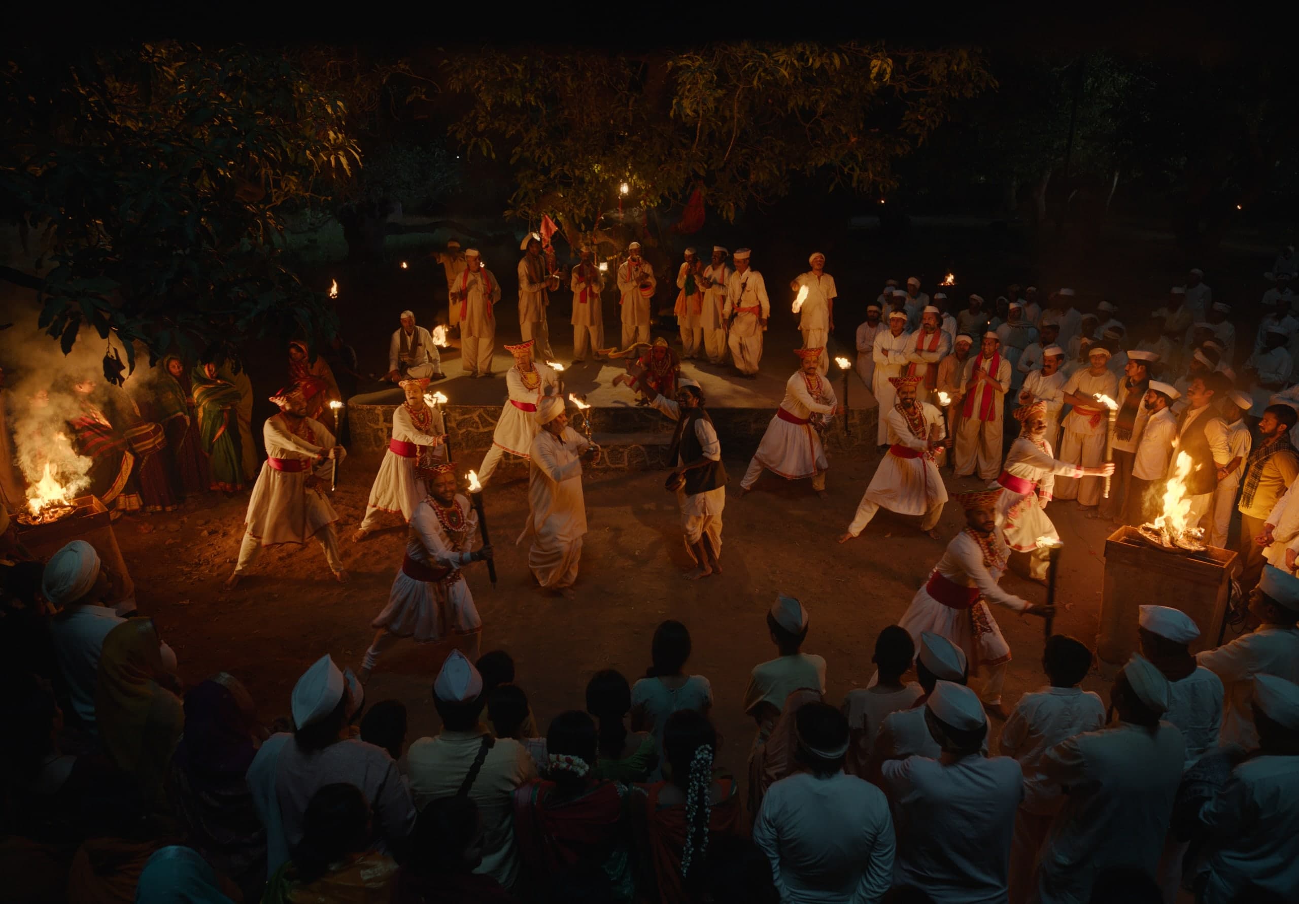 Seen from above at night, Gondhali performers in white and red dance with lit torches in front of a stone stage, surrounded by villagers.