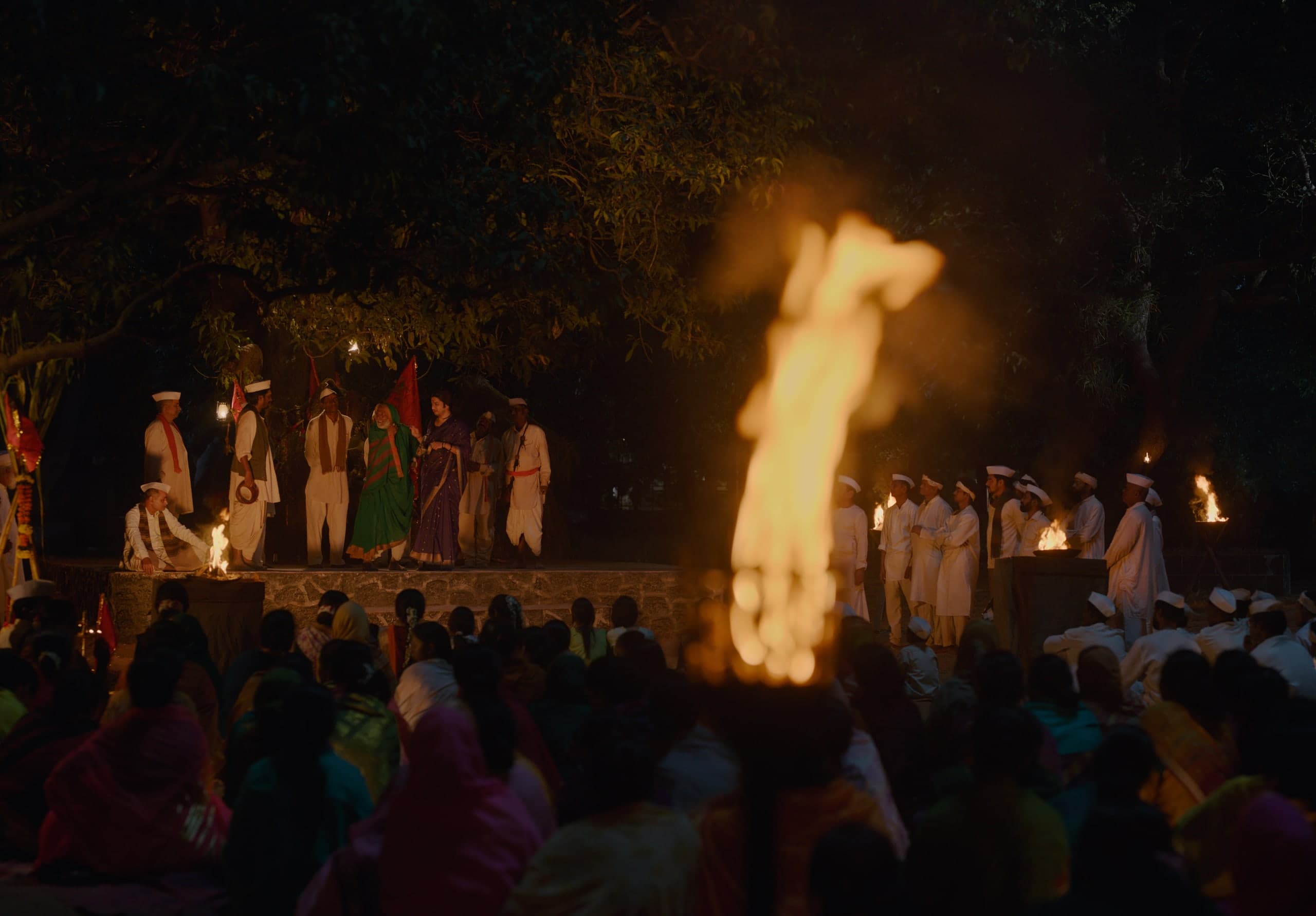 A large flame burns out of focus in the foreground; behind it, performers and red flags on a lit stone stage and a seated crowd in colourful saris.