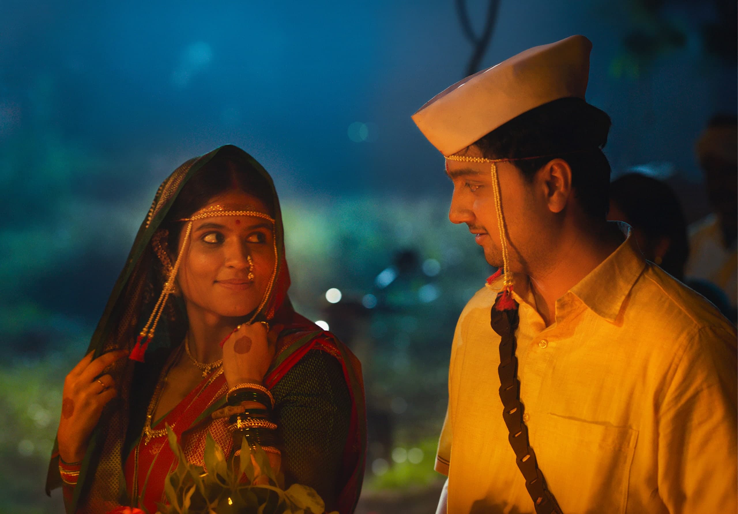 A bride in a red and green sari and a pearl mundavalya glances sideways at a groom in a yellow shirt and white cap, lit warmly against a blue night.