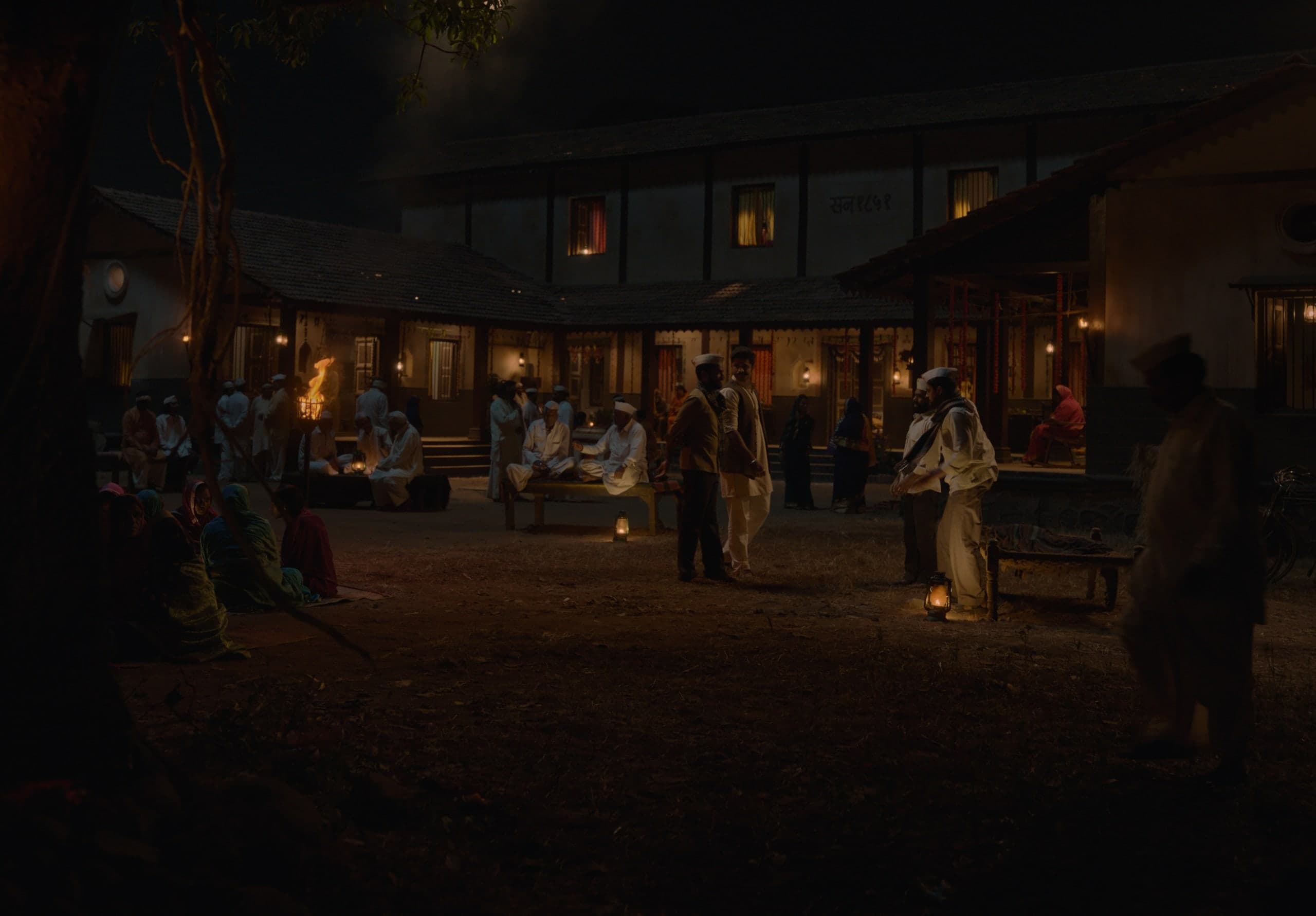 A two-storey village mansion with tiled roofs and lit windows at night; villagers gather in the courtyard around lanterns and a torch.