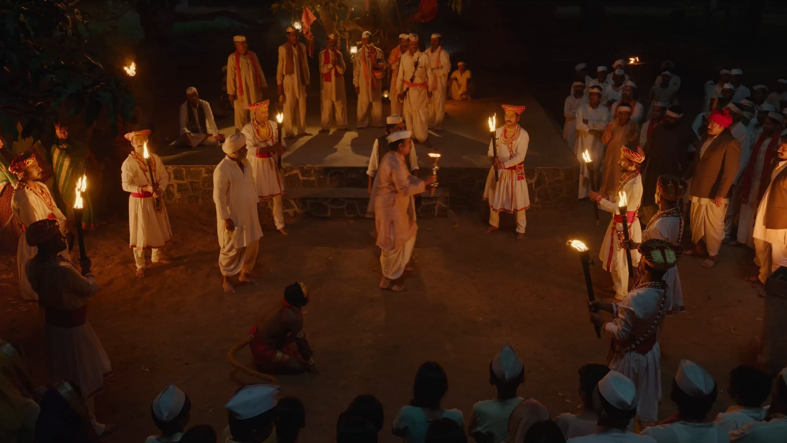 Seen from above at night, performers with lit torches dance in a ring before a stone stage as villagers watch.