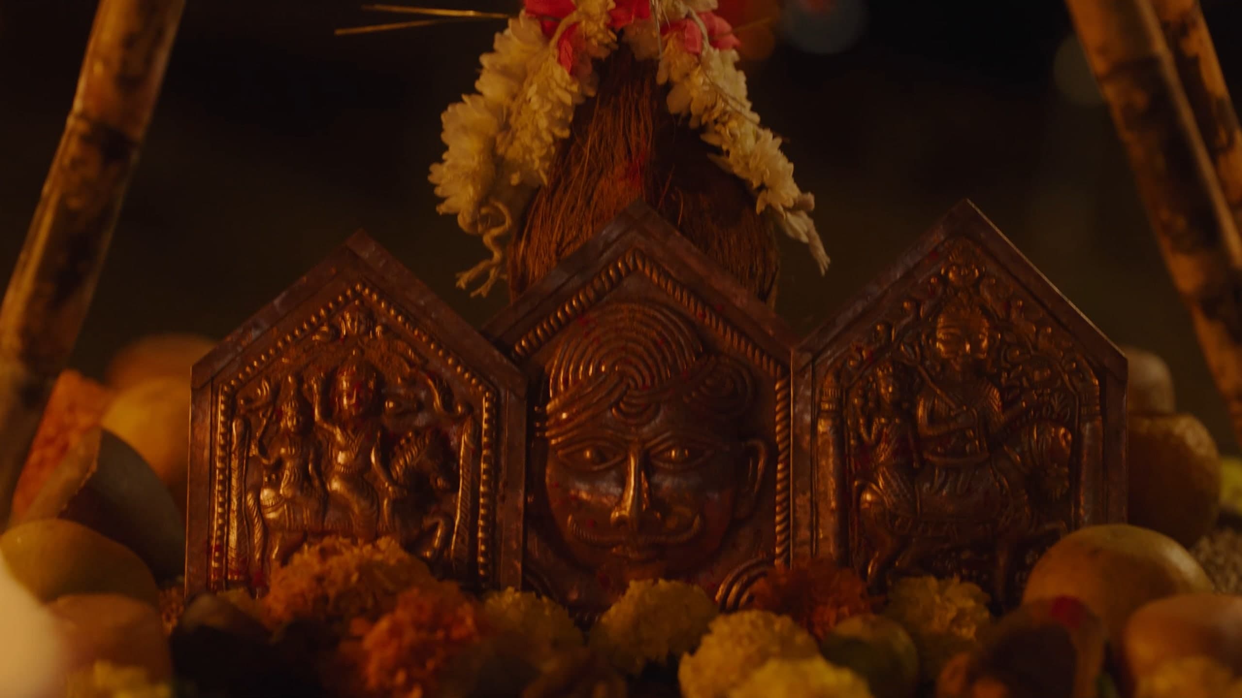A shrine at night: three embossed metal plaques, the centre one a face, decked with marigolds and a flower garland.